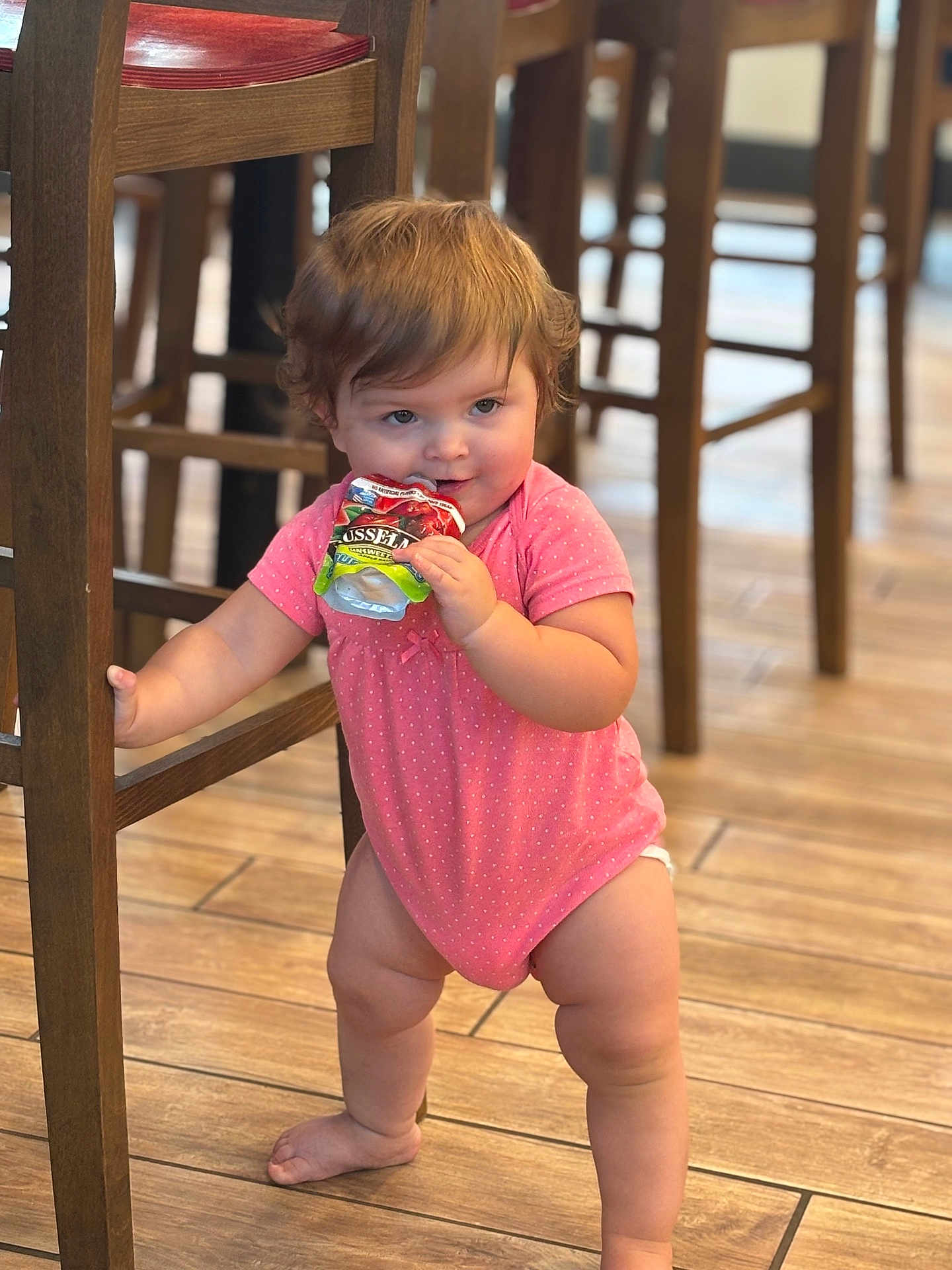 Amira joined the competition — help win amazing prizes! toddler, child, baby, pink_onesie, snack_pouch, wooden_chair, wooden_floor, indoor, standing, holding, cute, chubby_legs, polka_dot, furniture, flooring, young_child, candid, portrait, playful, expression
