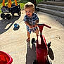 Thomas is registered to the contest to win money with this photo: toddler, child, toy_scooter, outdoor, sunlight, concrete, sidewalk_chalk, toy_car, playtime, happy, smiling, blue_shirt, barefoot, shadows, greenery, brick_wall, steps, play_area, daytime, summer