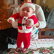 Emilien a rejoint le concours — aidez-le/la à gagner de superbes lots ! baby, adult, person, christmas, holiday, festive, santa_outfit, red_clothing, tablecloth, holly, ribbons, wooden_cabinet, christmas_tree, smile, indoor, holding, striped_socks, celebration, furniture, happy