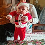 baby, adult, person, christmas, holiday, festive, santa_outfit, red_clothing, tablecloth, holly, ribbons, wooden_cabinet, christmas_tree, smile, indoor, holding, striped_socks, celebration, furniture, happy