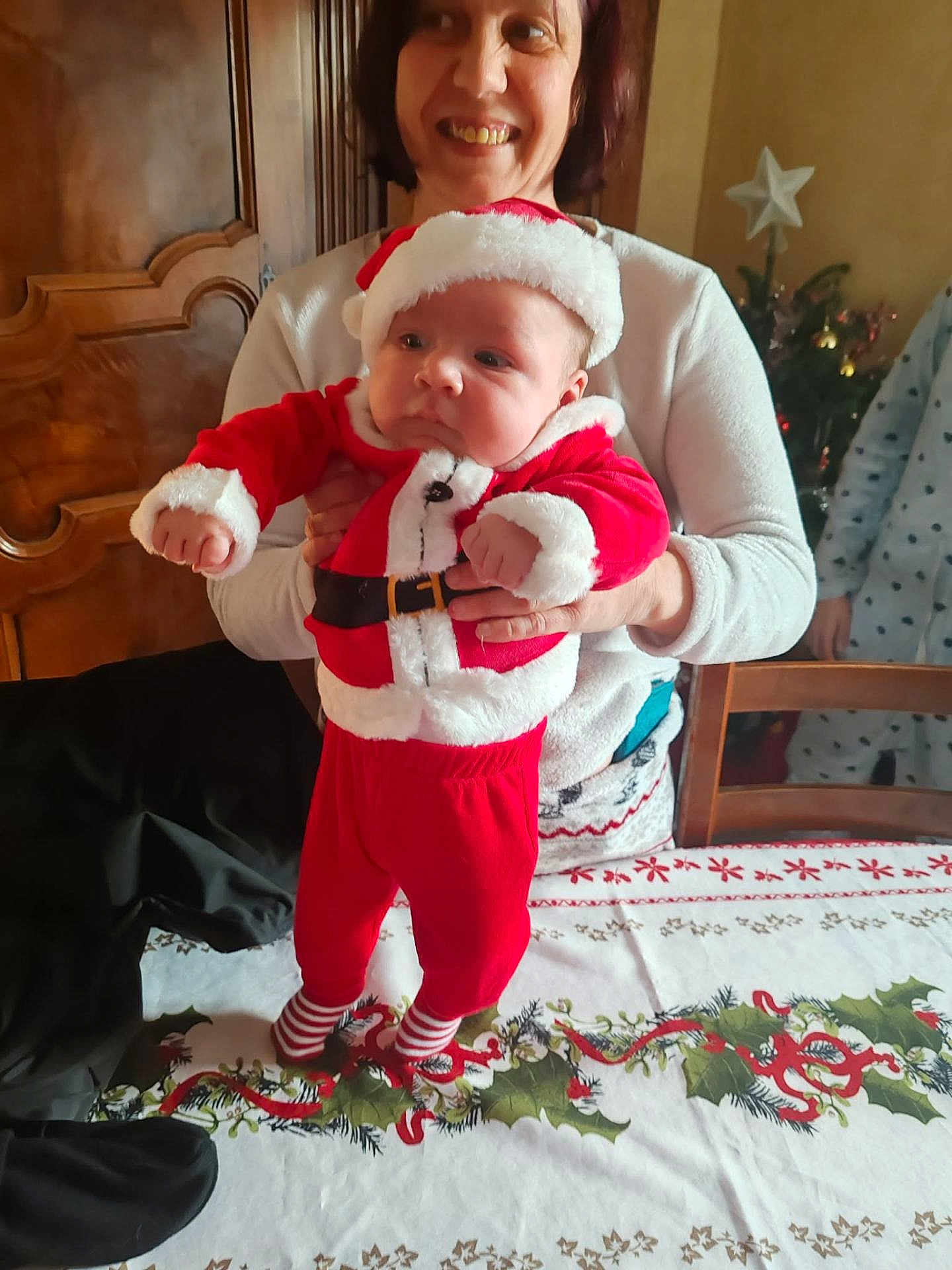 Emilien a rejoint le concours — aidez-le/la à gagner de superbes lots ! baby, adult, person, christmas, holiday, festive, santa_outfit, red_clothing, tablecloth, holly, ribbons, wooden_cabinet, christmas_tree, smile, indoor, holding, striped_socks, celebration, furniture, happy