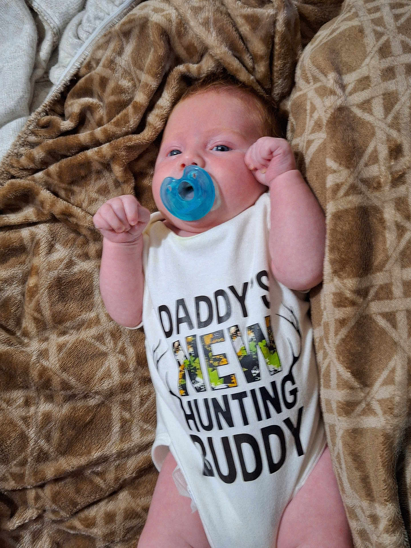 Noah is registered to the contest to win money with this photo: baby, infant, pacifier, onesie, blanket, lying_down, face, expression, cute, child, newborn, clothing, human, skin, hand, fist, blue, brown, pattern, portrait
