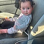 baby, child, car_seat, seat, clothing, socks, striped_socks, grey_shirt, curly_hair, rosy_cheeks, car_interior, seatbelt, blanket, sunlight, window, cup, floor, curious_expression, indoor, portrait