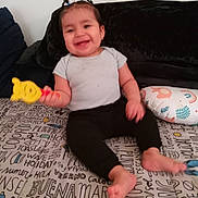 Michelle joined the competition — help win amazing prizes! toddler, child, baby, smiling, sitting, portrait, indoor, couch, bed, bedspread, toy, yellow_toy, hair_ties, black_pants, gray_shirt, barefoot, pillow, home, happy, playful