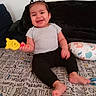 toddler, child, baby, smiling, sitting, portrait, indoor, couch, bed, bedspread, toy, yellow_toy, hair_ties, black_pants, gray_shirt, barefoot, pillow, home, happy, playful