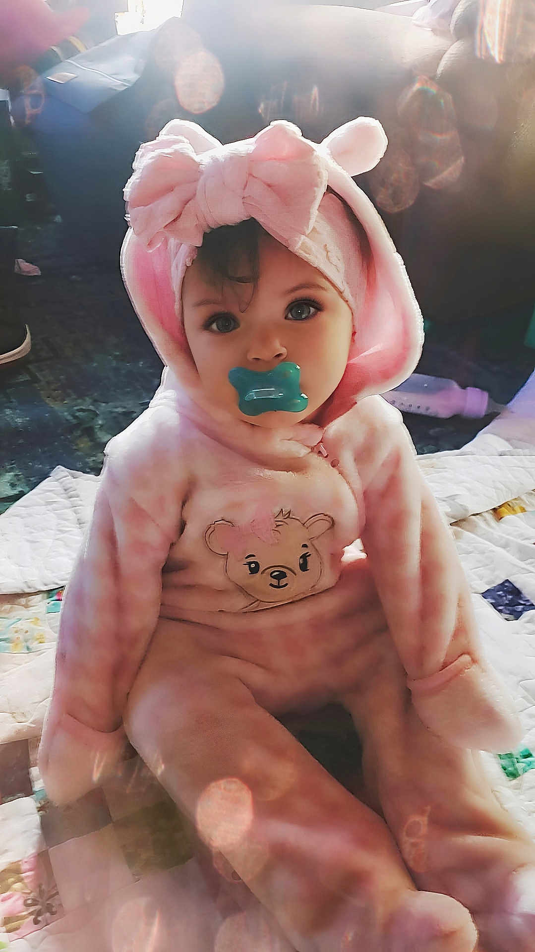 Ava joined the competition — help win amazing prizes! baby, child, pacifier, pink_clothing, onesie, headband, bow, bear_design, quilt, sitting, indoor, soft_light, cute, portrait, infant, face, eyes, cozy, plush, floor