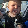 baby, child, infant, smiling, tongue_out, pacifier_clip, black_clothing, car_interior, sunlight, shadow, face, cheeks, short_hair, person, outdoor, window, seat, door, playful, cute