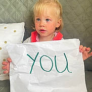 Mia participe au concours pour gagner de l'argent avec cette photo : child, toddler, blonde_hair, blue_eyes, paper, handwriting, couch, quilted, sitting, indoor, curious, expression, casual_clothing, holding, feet, pillow, home, furniture, face, person