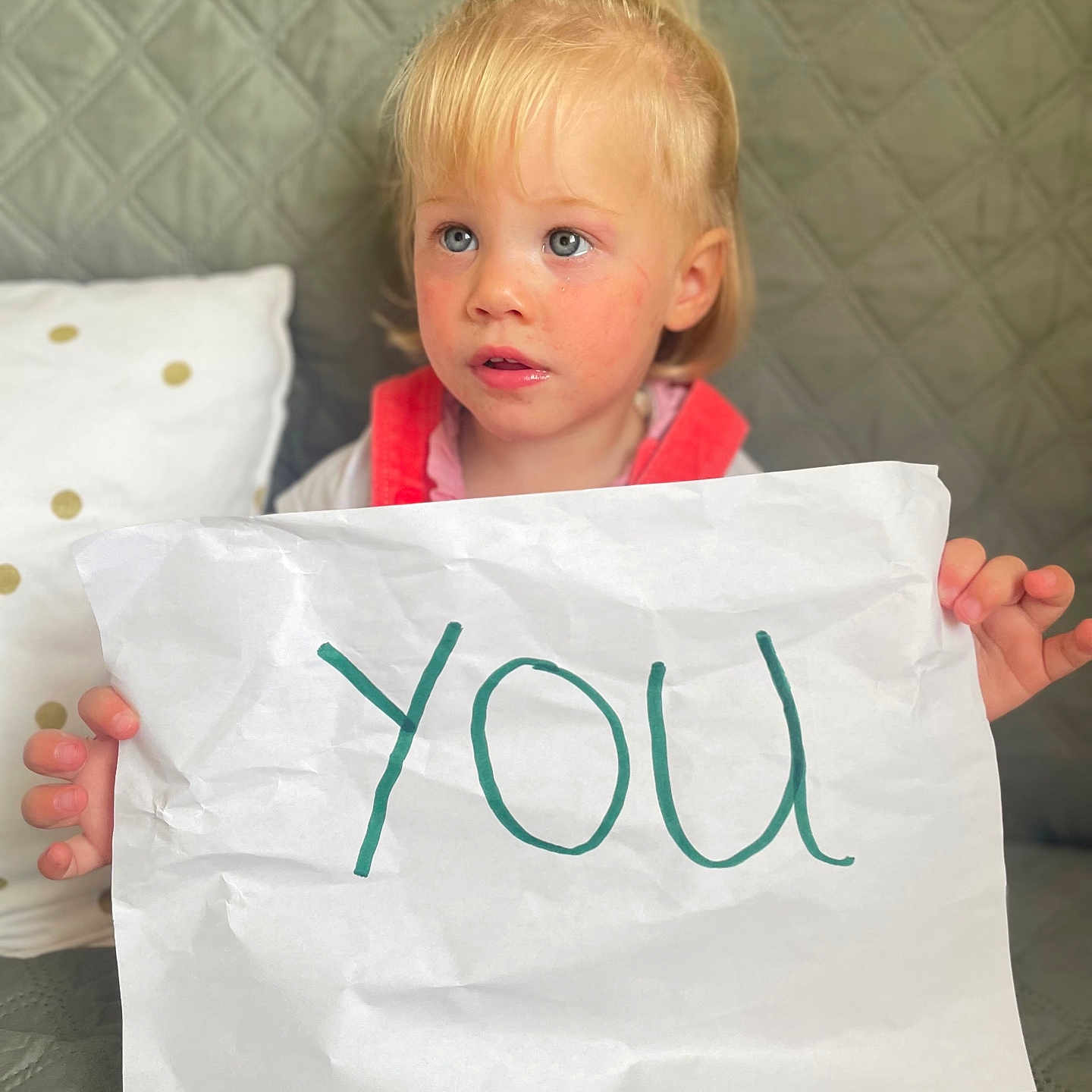 Mia participe au concours pour gagner de l'argent avec cette photo : blonde_hair, blue_eyes, casual_clothing, child, couch, curious, expression, face, feet, furniture, handwriting, holding, home, indoor, paper, person, pillow, quilted, sitting, toddler