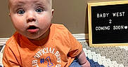 Bentley is registered to the contest to win money with this photo: baby, child, infant, orange_shirt, blue_eyes, crocheted_blanket, ultrasound_photos, letter_board, announcement, surprised_expression, sitting, indoor, casual_clothing, family, pregnancy_announcement, cute, portrait, young_child, home, floor