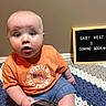 baby, child, infant, orange_shirt, blue_eyes, crocheted_blanket, ultrasound_photos, letter_board, announcement, surprised_expression, sitting, indoor, casual_clothing, family, pregnancy_announcement, cute, portrait, young_child, home, floor