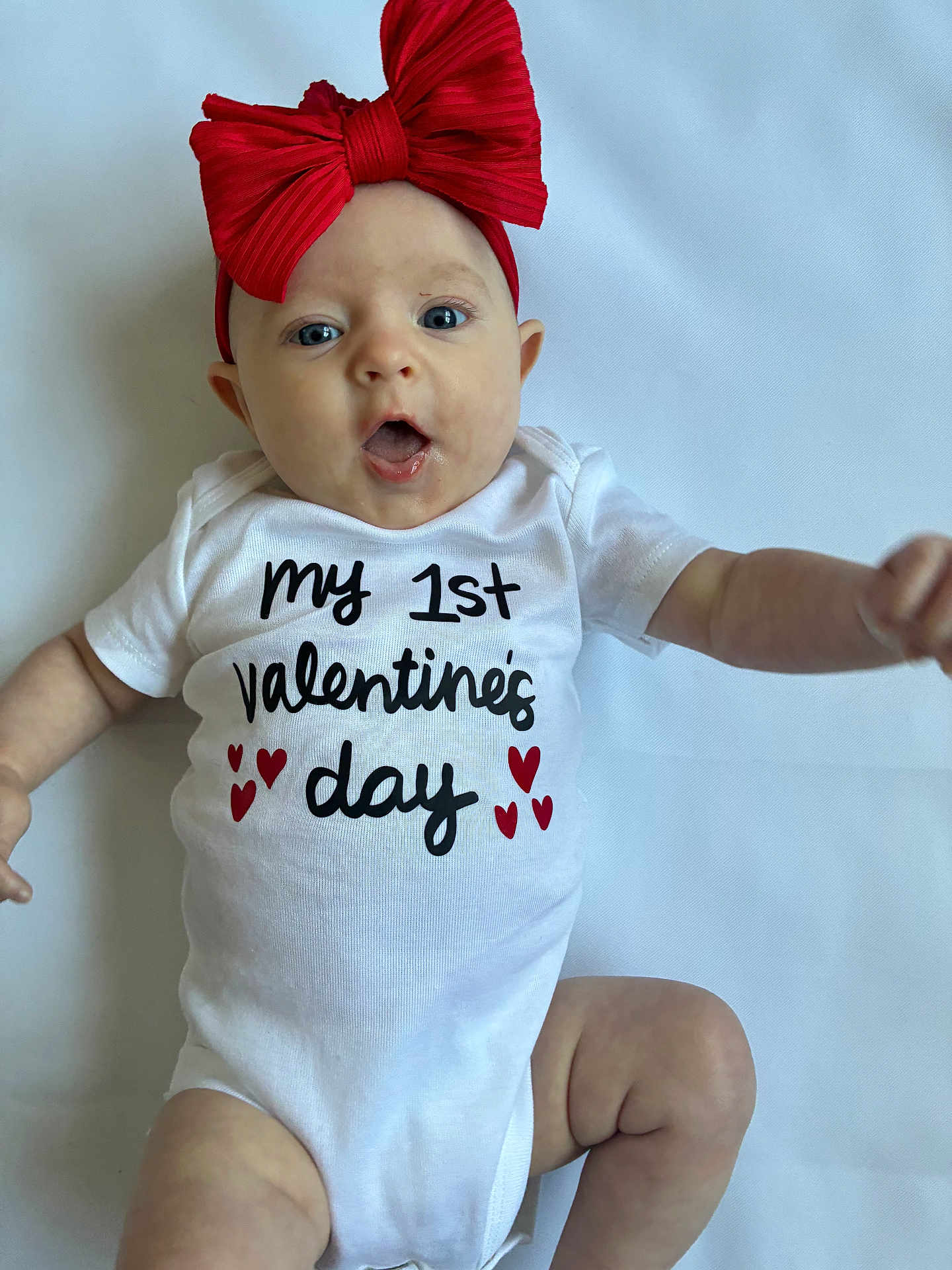 Hadleigh joined the competition — help win amazing prizes! child, baby, infant, onesie, bow, red_bow, valentine, hearts, face, eyes, mouth, surprised_expression, lying_down, blanket, white_background, cute, clothing, portrait, hand, leg