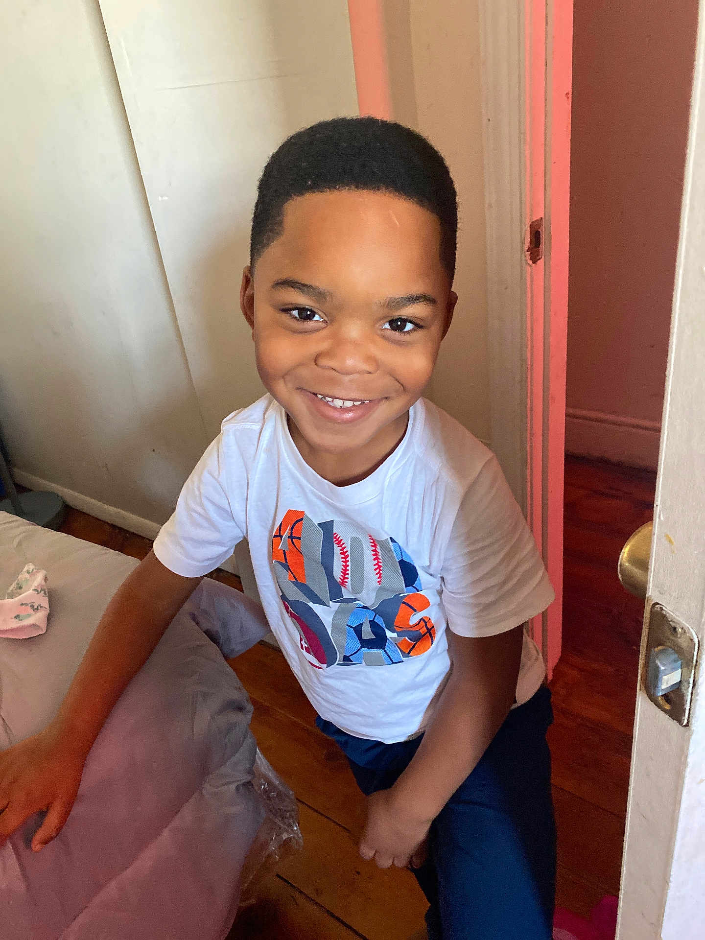 Carter is registered to the contest to win money with this photo: bedroom, boy, casual_clothing, child, door_handle, doorway, eyes, hand, happy, hardwood_floor, indoor, mattress, person, portrait, short_hair, smile, sports_graphic, teeth, wall, white_tshirt