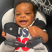 Amir is registered to the contest to win money with this photo: baby, infant, smile, car_seat, harness, buckle, push_label, onesie, pacifier_clip, curly_hair, brown_eyes, hand, closeup, portrait, fabric_pattern, seat_padding, cute, happy, safety_strap, indoor