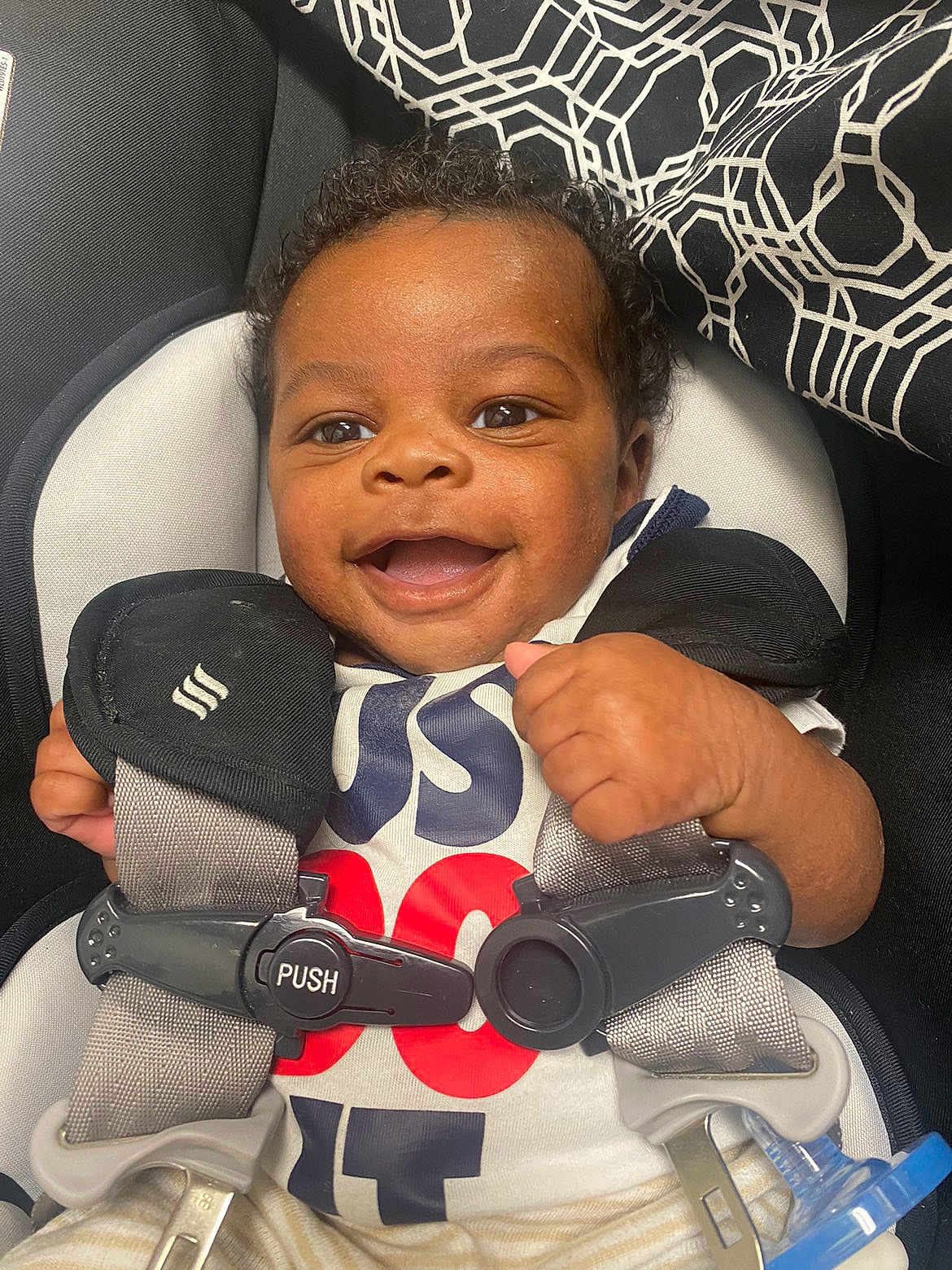 Amir is registered to the contest to win money with this photo: baby, infant, smile, car_seat, harness, buckle, push_label, onesie, pacifier_clip, curly_hair, brown_eyes, hand, closeup, portrait, fabric_pattern, seat_padding, cute, happy, safety_strap, indoor