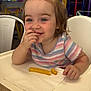Malee is registered to the contest to win money with this photo: child, toddler, high_chair, french_fries, eating, smiling, blue_eyes, pigtails, hair_clips, striped_shirt, napkin, playground, indoor, colorful_background, table, hand, portrait, food, happy, candid