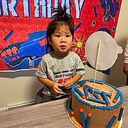 Caius joined the competition — help win amazing prizes! child, toddler, birthday, cake, table, banner, toy_gun, foam_darts, cake_decoration, hair_tie, curious, indoor, party, celebration, person, clothing, gray_shirt, orange, blue, face