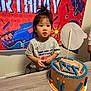 child, toddler, birthday, cake, table, banner, toy_gun, foam_darts, cake_decoration, hair_tie, curious, indoor, party, celebration, person, clothing, gray_shirt, orange, blue, face