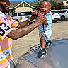 toddler, man, smiling, tattoos, car, hood, blue_sky, parking_lot, outdoor, holding_hands, child, shorts, sneakers, cap, happy, person, daylight, casual_clothing, buildings, vehicles