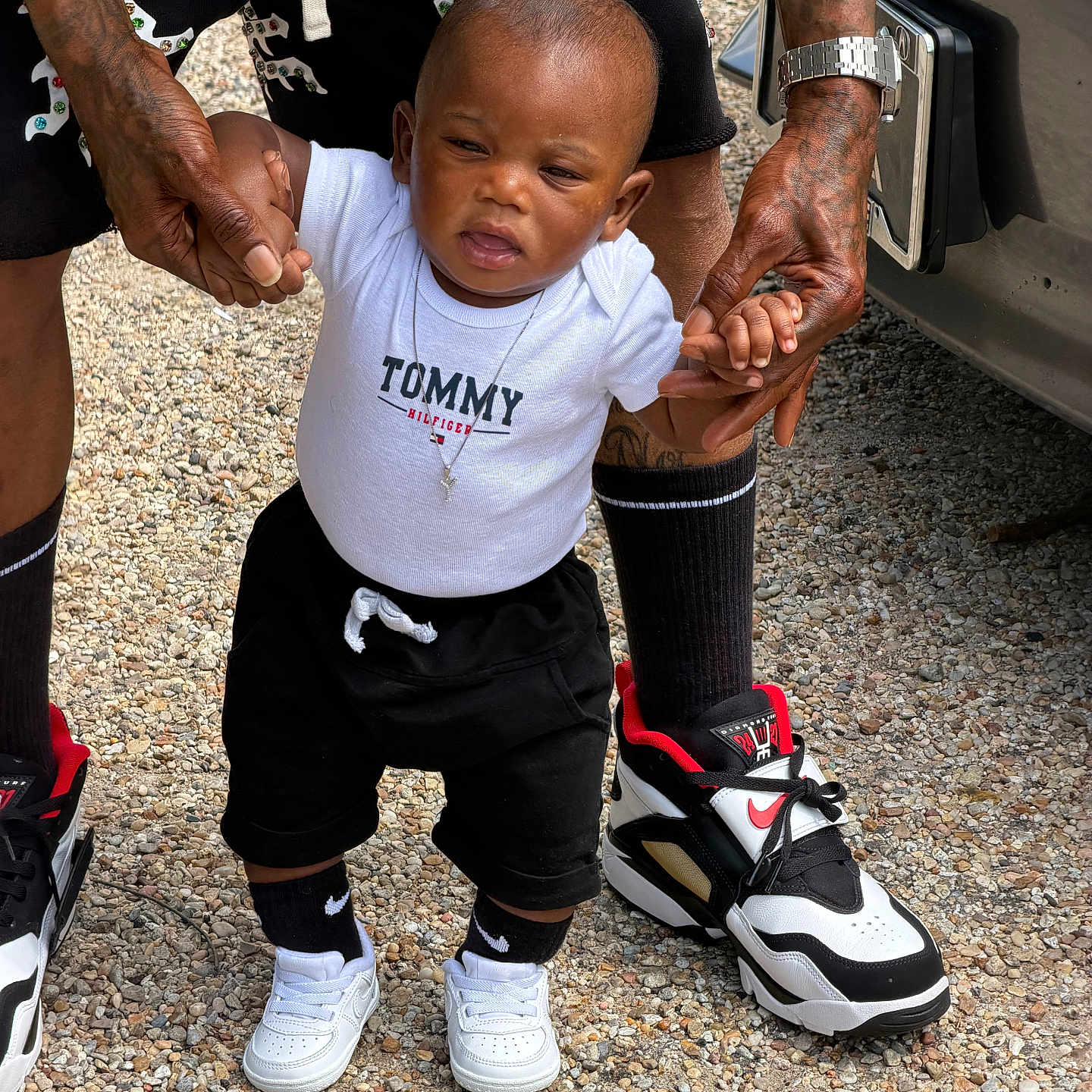 Yasir is registered to the contest to win money with this photo: toddler, child, adult, hands, tattoos, white_shirt, black_shorts, sneakers, gravel, outdoor, walking, support, person, fashion, footwear, standing, young_child, casual_clothing, daylight, holding_hands