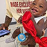 Yasir is registered to the contest to win money with this photo: baby, smile, red_bow, bottle, tattoo, smartphone, adult, jeans, watch, sneakers, white_shirt, text_on_shirt, indoor, tile_floor, happy, child, person, clothing, hand, portrait