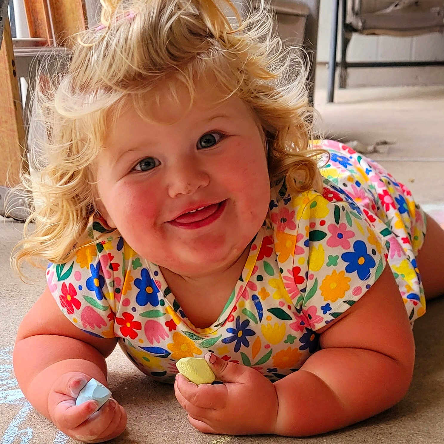 Millie-Jean is registered to the contest to win money with this photo: child, toddler, blonde_hair, curly_hair, chalk, colorful, floral_shirt, smiling, rosy_cheeks, floor, drawing, playful, happy, indoor, cute, person, hands, face, cheeks, casual_clothing