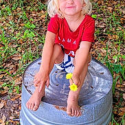 Jordan-Lee is registered to the contest to win money with this photo: child, toddler, blond_hair, red_shirt, shorts, barefoot, flower, metal_tub, outdoor, grass, leaves, fence, nature, playful, smiling, squatting, summer, greenery, cute, happy