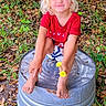 child, toddler, blond_hair, red_shirt, shorts, barefoot, flower, metal_tub, outdoor, grass, leaves, fence, nature, playful, smiling, squatting, summer, greenery, cute, happy