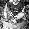 child, blond_hair, sitting, barefoot, metal_barrel, outdoor, fence, leaves, black_and_white, tshirt, shorts, portrait, young_child, curly_hair, nature, casual, playful, expression, grass, yard