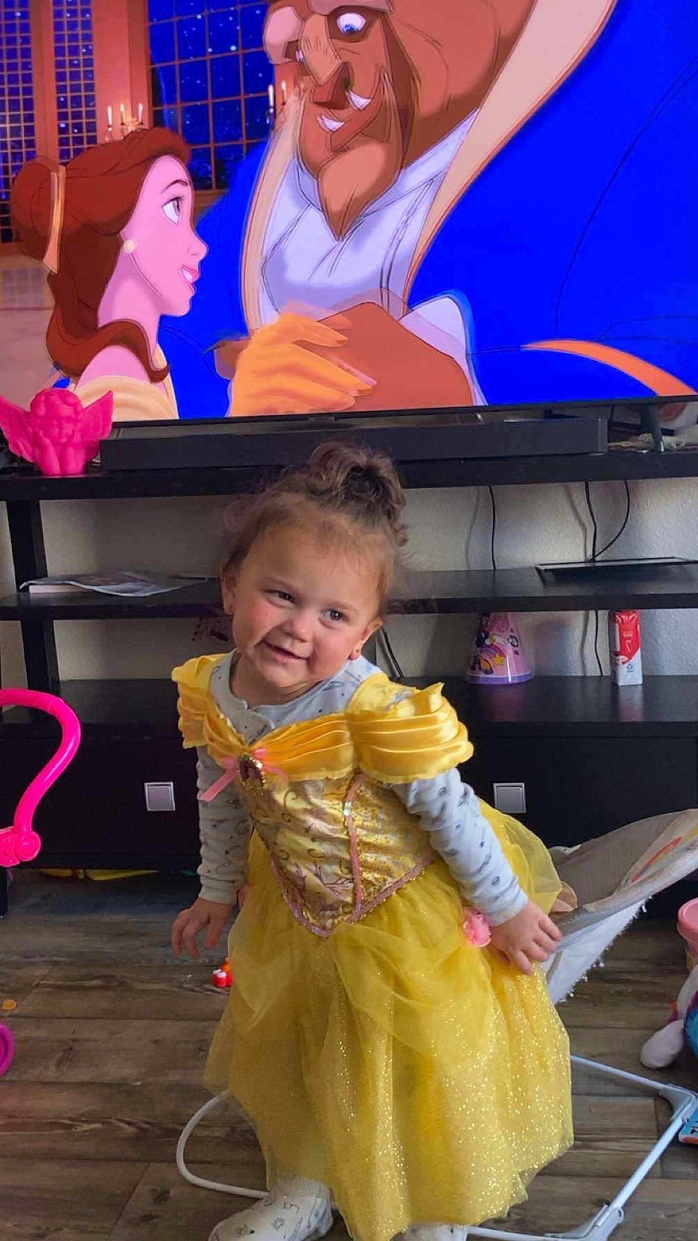 Valerie is registered to the contest to win money with this photo: child, toddler, yellow_dress, princess_costume, smiling, indoor, television, animated_movie, beauty_and_the_beast, toy, floor, furniture, living_room, curly_hair, playful, cute, person, sock, chair, entertainment
