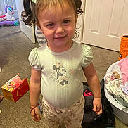 Valerie is registered to the contest to win money with this photo: baby_items, bows, carpet, child, door, floor, girl, home, indoor, laundry_basket, minnie_mouse, pants, person, pigtails, plastic_toy, shirt, smile, socks, toddler, toys