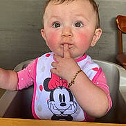 Athena is registered to the contest to win money with this photo: baby, child, high_chair, pink_bow, minnie_mouse, bib, finger_in_mouth, rosy_cheeks, bracelet, curious, indoor, restaurant, table, wooden_chair, cute, portrait, expression, young_child, person, casual_clothing