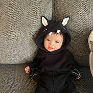 Emiliana is registered to the contest to win money with this photo: baby, child, hoodie, black_clothing, ears, teeth, smile, couch, gray, texture, sitting, infant, cute, clothing, portrait, indoor, person, young_child, face, happy