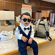 George is registered to the contest to win money with this photo: toddler, child, sunglasses, bow_tie, suit, kitchen, counter, backpack, hands, wood_paneling, cabinet, coffee_maker, microwave, plastic_bag, person, indoor, table, shoes, cool, portrait