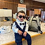 toddler, child, sunglasses, bow_tie, suit, kitchen, counter, backpack, hands, wood_paneling, cabinet, coffee_maker, microwave, plastic_bag, person, indoor, table, shoes, cool, portrait