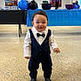 toddler, child, boy, formal_clothing, tuxedo, bow_tie, standing, smiling, indoor, balloons, party, floor, table, blue, black, poster, person, celebration, cute, happy