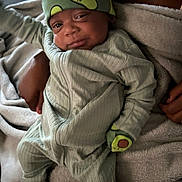 Travis is registered to the contest to win money with this photo: baby, infant, avocado_hat, pajamas, mittens, blanket, soft_texture, smile, sleepy, comfort, child, cute, cozy, hands, footwear, portrait, indoors, warm, relaxed, person