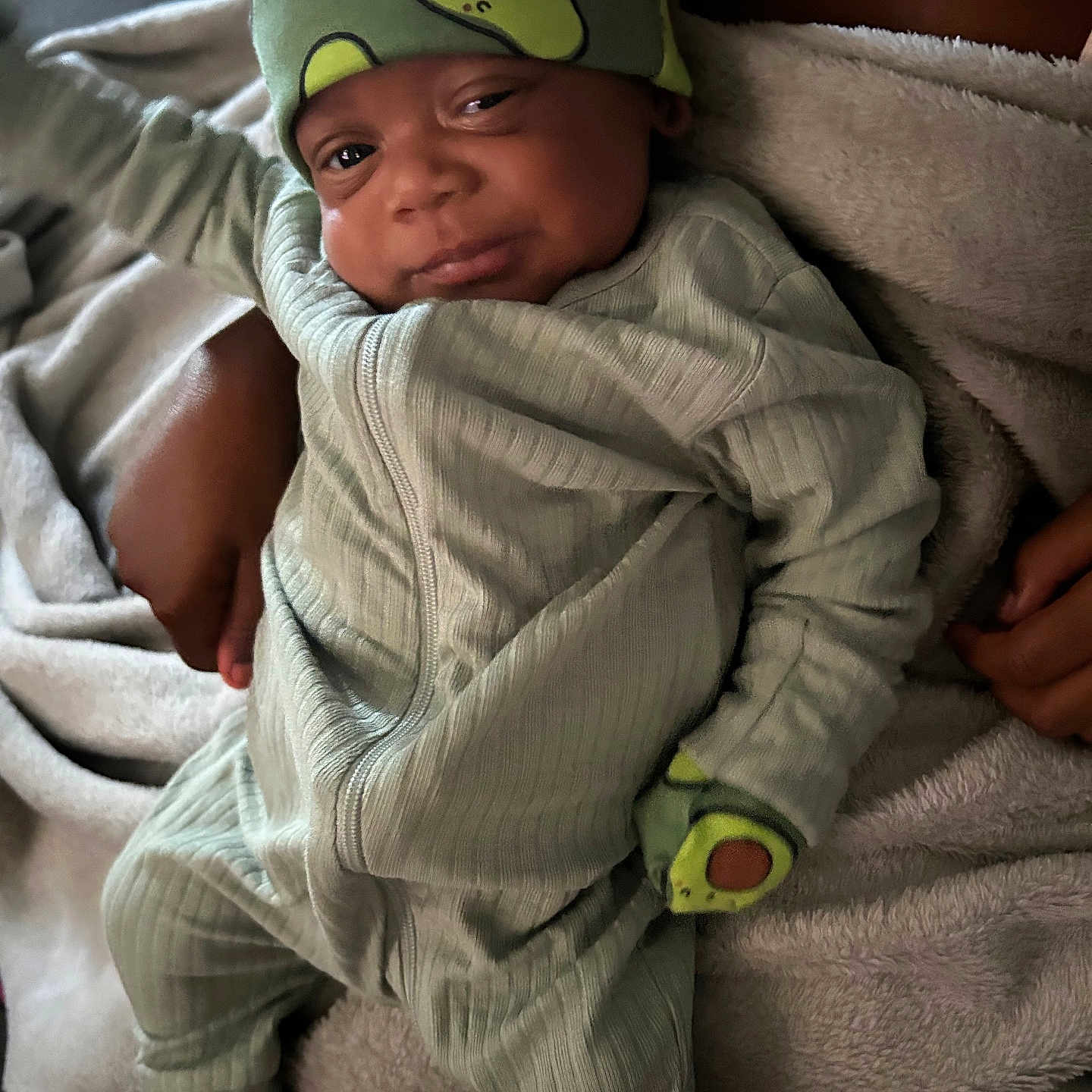Travis is registered to the contest to win money with this photo: avocado_hat, baby, blanket, child, comfort, cozy, cute, footwear, hands, indoors, infant, mittens, pajamas, person, portrait, relaxed, sleepy, smile, soft_texture, warm