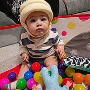 Iaith joined the competition — help win amazing prizes! baby, child, helmet, ball_pit, colorful_balls, toy, indoor, sitting, couch, hand, striped_shirt, socks, plush_toy, person, face, expression, playtime, soft_lighting, home, curious