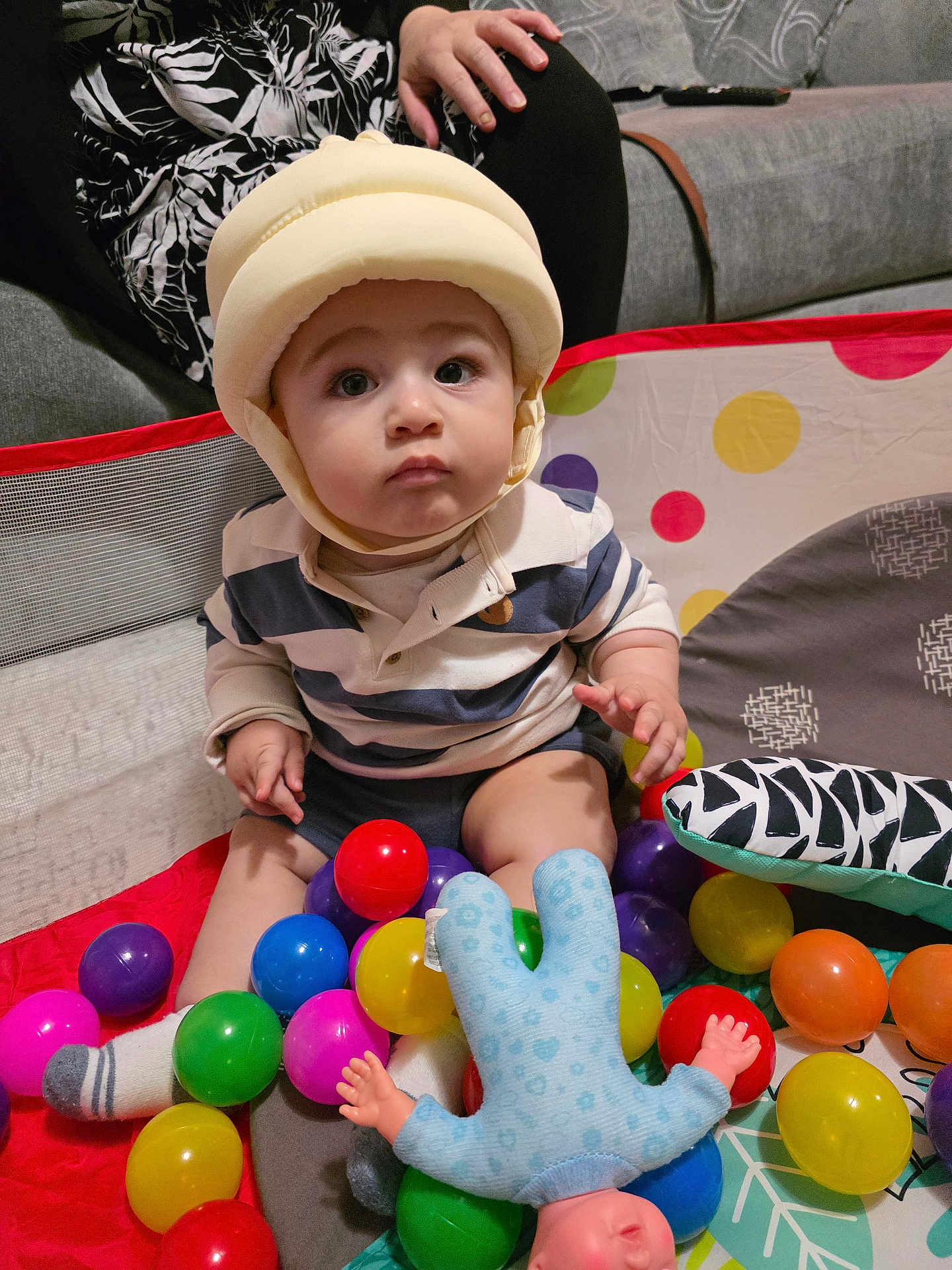 Iaith joined the competition — help win amazing prizes! baby, child, helmet, ball_pit, colorful_balls, toy, indoor, sitting, couch, hand, striped_shirt, socks, plush_toy, person, face, expression, playtime, soft_lighting, home, curious