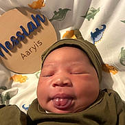 Nessiah joined the competition — help win amazing prizes! baby, blanket, child, clothing, cozy, cute, dinosaur_pattern, face, hat, indoors, infant, name_plaque, newborn, portrait, sleepy, smiling, soft_light, tongue_out, wooden_sign, wrapped