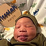 baby, blanket, child, clothing, cozy, cute, dinosaur_pattern, face, hat, indoors, infant, name_plaque, newborn, portrait, sleepy, smiling, soft_light, tongue_out, wooden_sign, wrapped