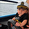baby, captain_hat, boat, steering_wheel, water, lake, cloudy_sky, child, person, outdoor, black_shirt, hand, face, young_child, nautical, transportation, summer, cute, portrait, expression