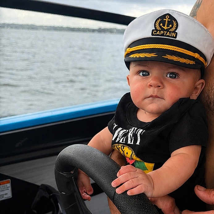 Brantley Tharpe is registered to the contest to win money with this photo: baby, captain_hat, boat, steering_wheel, water, lake, cloudy_sky, child, person, outdoor, black_shirt, hand, face, young_child, nautical, transportation, summer, cute, portrait, expression