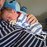 baby, newborn, sleeping, pacifier, striped_clothing, adult, hand, plush_toy, blue_toy, indoor, close_up, comfort, rest, soft_lighting, cute, child, person, casual, relaxation, care