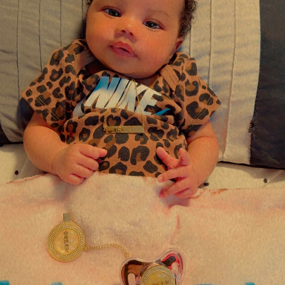 Dream is registered to the contest to win money with this photo: accessory, baby, bed, blanket, child, clothing, curly_hair, cute, face, gold_necklace, human, indoors, infant, leopard_print, lying_down, nike_shirt, pacifier, portrait, skin, soft_lighting