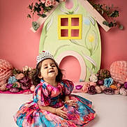 Tiana is registered to the contest to win money with this photo: toddler, child, girl, dress, tiara, flower, floral_dress, smiling, sitting, indoor, portrait, colorful, cute, happy, decor, pink_background, playful, young_child, innocence, celebration