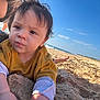 toddler, child, beach, sand, sky, ocean, waves, mustard_sweater, curly_hair, face, expression, sunny, outdoor, playful, closeup, casual_clothing, barefoot, nature, portrait, daytime