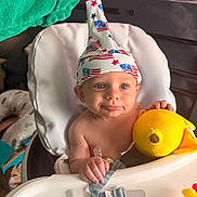 Brian is registered to the contest to win money with this photo: baby, child, high_chair, plush_toy, hat, american_flag, hand, smiling, indoor, face, person, cute, bright_eyes, clothing, furniture, toy, white, green, yellow, happy