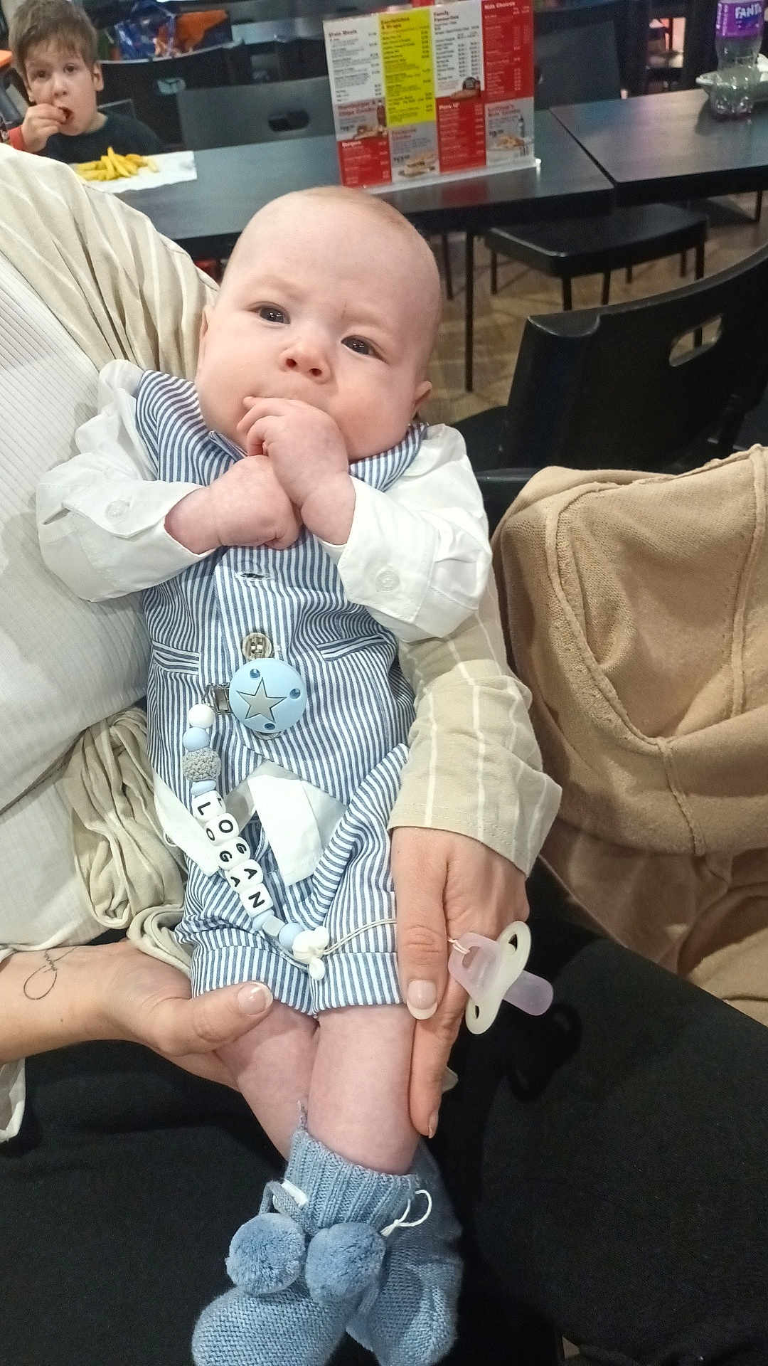 Logan is registered to the contest to win money with this photo: baby, child, person, infant, holding, clothing, blue_booties, pacifier, name_tag, restaurant, table, food, fries, young_child, hands, seated, casual, indoor, family, cute